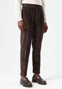 Pantalon en velours côtelé marron à taille haute, avec plis à l'avant et revers retroussés. Associé à des mocassins marron foncé. Surface texturée visible.