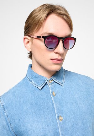 Giovane uomo con capelli chiari indossa occhiali da sole rotondi con lenti sfumate rosse e una camicia di jeans azzurro chiaro con bottoni.