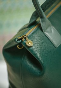 Green leather handbag featuring a textured finish, gold zipper, and a round zipper pull with embossed detailing. Robust handles complete the design.