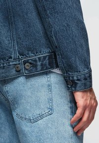 Giacca di denim blu scuro, con tessuto testurizzato, dettagli con bottoni e orlo leggermente sfilacciato, abbinata a jeans azzurri.