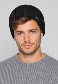 Schwarze, gerippte Mütze aus weichem Stoff. Verfügt über eine runde Krone und dezentes Branding an der Seite. Wird mit einem grauen, gerippten Pullover getragen.