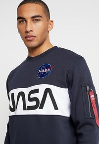 Sudadera navy con una banda blanca que presenta "NASA" en letras negras en negrita, logo de NASA bordado en el pecho y un bolsillo con cremallera con acentos rojos.