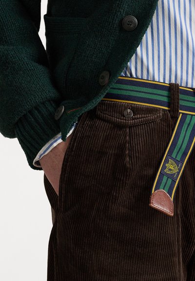 Sötétbarna kordbársony nadrág zöld, kék és sárga csíkos övvel; zöld kötött kardigán; kék és fehér csíkos ing alatt.