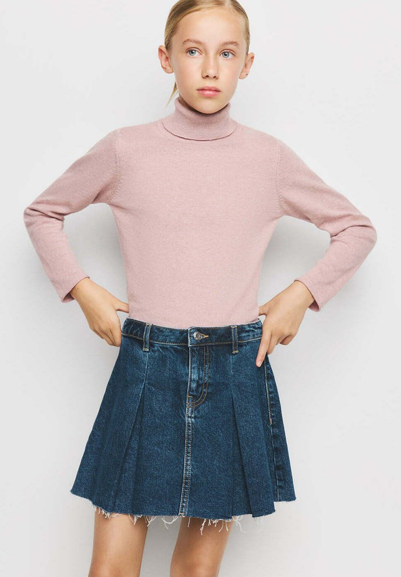 Jeune personne aux cheveux blonds portant un pull à col roulé rose et une jupe plissée en denim bleu foncé, debout les mains sur les hanches.