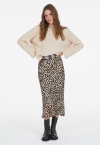 Beige gestrickter Pullover mit weitem Ärmel, kombiniert mit einem midi Rock in Leopardenmuster, der eine figurbetonte hohe Taille und eine gerade Silhouette hat. Schwarze Stiefeletten.
