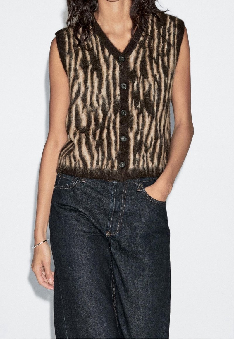 Femme portant un gilet sans manches en tissu duveteux marron et beige à motifs avec des boutons noirs et un jean en denim bleu foncé, une main dans la poche.