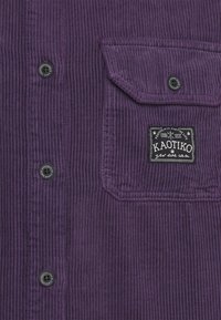 Camisa de pana morada con un bolsillo con un botón negro y una etiqueta de logo blanca, que muestra una textura de rayas verticales.