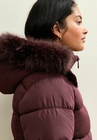 Chaqueta acolchada burdeos con capucha forrada de piel, que cuenta con un cierre de cremallera y un diseño acolchado voluminoso. Mangas texturizadas y detalles de costura sutiles.
