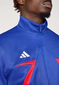Blå atletisk zip-up jakke laget av lett stoff, med høy krage, røde detaljer og en hvit Adidas-logo på brystet.