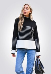 Schwarz-weißer gestreifter Pullover mit hohem Kragen und Rippstruktur, kombiniert mit einer hellblauen Jeans und einer schwarzen Tasche.