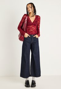 Top de renda burgundy de manga longa com decote ondulado, combinado com jeans escuros de perna larga. Acessórios incluem uma bolsa vermelha e sapatos pretos.