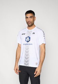 Λευκό αθλητικό κοντομάνικο μπλουζάκι με μαύρες λεπτομέρειες, σχέδιο φιδιού και λογότυπο. Συνδυάζεται με μαύρο παντελόνι που φέρει πορτοκαλί λογότυπο.