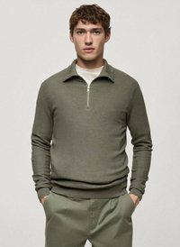 Pullover verde olivo, con texture, colletto con zip, maniche lunghe e design aderente, abbinato a pantaloni grigi chiari.