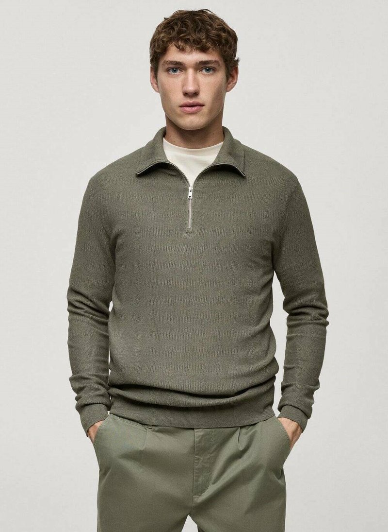 Pullover verde olivo, con texture, colletto con zip, maniche lunghe e design aderente, abbinato a pantaloni grigi chiari.