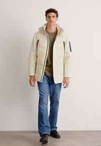 Daunenjacke in Creme mit Kapuze und schwarzen Reißverschlüssen. Über einem olivgrünen Pullover getragen, kombiniert mit Jeans und schwarzen Schuhen.