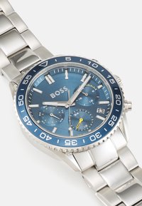 Montre en acier inoxydable avec un cadran bleu, des aiguilles lumineuses, un affichage de la date et des accents jaunes. La lunette comporte des marquages blancs et une finition texturée.