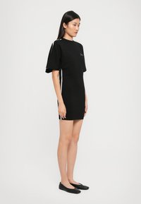 Γυναίκα που στέκεται στο πλάι, ντυμένη με ένα κοντό μαύρο φόρεμα με λευκές ρίγες στο πλάι και μαύρες φλατ σόλες, σε απλό λευκό φόντο.