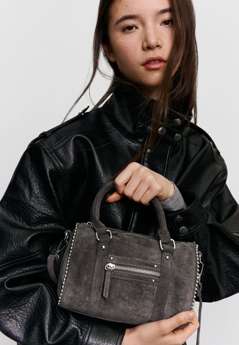 Jeune femme en veste en cuir noire tenant un petit sac à main en daim gris avec fermeture éclair et clous argentés sur un fond uni.