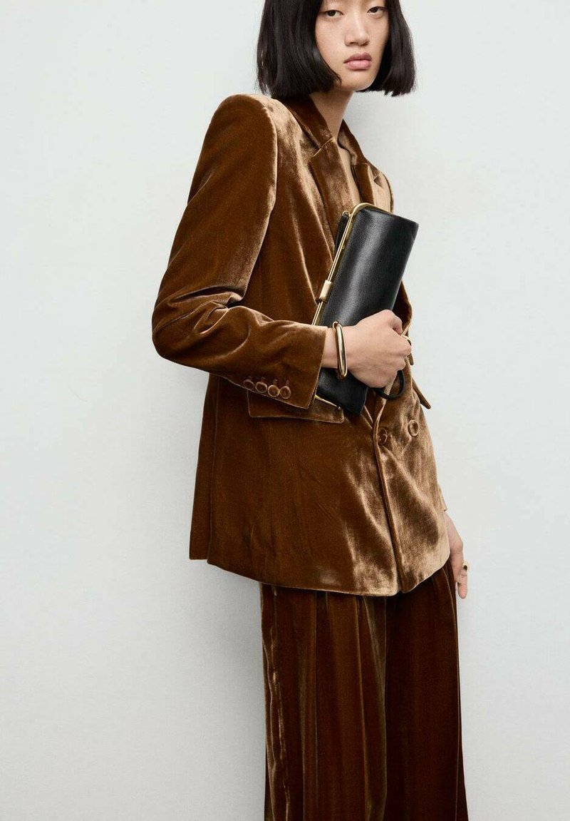 Blazer in velluto marrone con pantaloni coordinati, caratterizzato da un rever a ragto e un leggero luccichio. Il modello tiene una pochette nera con un accento dorato.