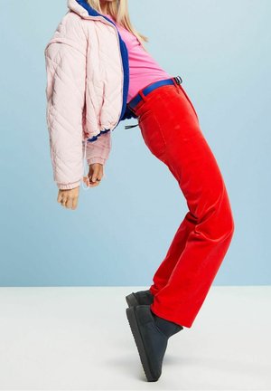 Vadderad rosa jacka med blått foder, tajta röda manchesterbyxor och svarta platåskor. Modell poserar med lutande hållning mot en blå bakgrund.