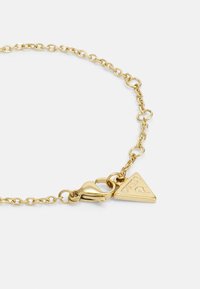 Gold metal bracelet with a chain link design, featuring a triangular charm that is engraved with the word "GUESS" and a clasp fastening.