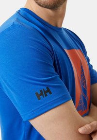 Camiseta de manga corta azul hecha de tejido suave, con un diseño gráfico naranja y un logo negro en la manga izquierda.