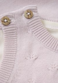 Ljust rosa stickad plagg med en ribbad halsringning, två runda knappar och texturerade blommönster på tyget. Mjuk inre layer synlig.