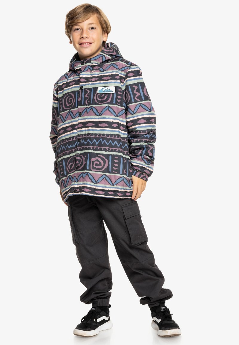 Chaqueta con capucha colorida y estampada con diseños en espiral y geométricos en púrpura, negro y azul. Combinada con pantalones cargo negros y zapatillas.
