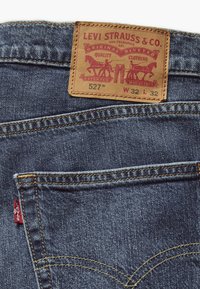Vaqueros de mezclilla con un parche de cuero marrón que presenta letras rojas y un logo, un hilo amarillo en las costuras y una pequeña etiqueta roja en el bolsillo trasero.