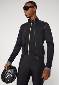 Veste de cyclisme noire au design ajusté, avec des accents réfléchissants et une ouverture zippée, accompagnée de shorts de cyclisme noirs et d'un casque.