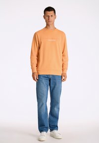 Felpa arancione con scollo a giro e testo stampato, abbinata a jeans blu a gamba dritta e sneakers bianche. Texture liscia in tutto il capo.