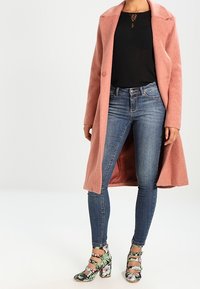 Rosa, langer Wollmantel über einem schwarzen Oberteil, kombiniert mit blauen Skinny-Jeans und hochhackigen Schuhen mit Blumenmuster und Knöchelriemen.