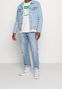 Ljusblå denimjacka över en vit t-shirt med grön grafik, kombinerat med ljusblå jeans och vita sneakers. Avslappnad outfit.