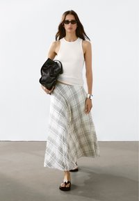 Débardeur blanc à côtes, jupe longue à carreaux crème et noir, sandales noires épaisses, et sac noir à texture douce. Bracelet manchette en argent.