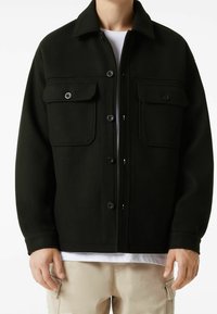 Černá bunda na knoflíky s límcem, dvěma náprsními kapsami a texturovanou tkaninou. Nosená přes bílou košili, spárovaná s béžovými cargo kalhotami.