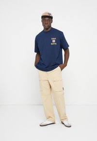 Marineblå bomulds t-shirt med "Carhartt University" grafik, parret med lyse beige cargo bukser med sidelommer og en kulgrå label. Hvide sneakers.