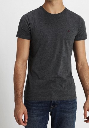 Mężczyzna ubrany w dopasowany, ciemnoszary, krótki rękaw T-shirt i niebieskie dżinsy na jednolitym jasnym tle.