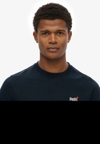 Sudadera azul marino con cuello redondo, hecha de algodón, que presenta un pequeño logo blanco y rojo en el lado izquierdo del pecho, con una textura suave.
