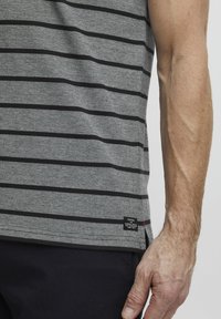 Grå ermeløs skjorte med svarte horisontale striper, med en liten merkeetikett på kanten og et teksturert stoff.