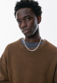 Bruine geribbelde gebreide trui met een ronde hals, die over een grijze shirt is gedragen, verfraaid met een zilveren kettinghalsketting.