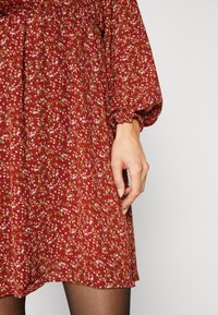 Vestido floral rojo con pequeñas flores rosas y blancas sobre una tela texturizada. Mangas largas abullonadas con puños elásticos y cintura fruncida.