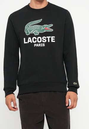 Czarna bluza z kapturem z zielonym logo krokodyla i napisem „LACOSTE PARIS”, noszona przez osobę w ciemnych spodniach.