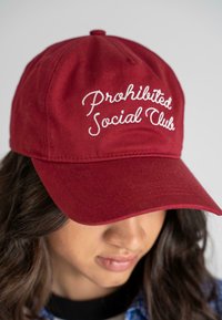Căciulă de baseball din bumbac roșu, cu text brodat "Prohibited Social Club" în alb, având o margine curbată și o siluetă structurată.