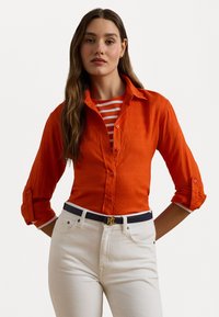 Orange linen button-up shirt with rolled sleeves layered over a red and white striped top, paired with white jeans and a navy belt with a gold logo.