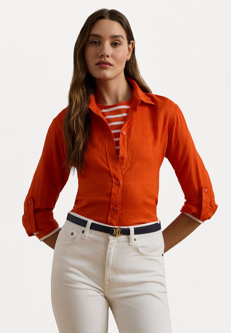 Orange linen button-up shirt with rolled sleeves layered over a red and white striped top, paired with white jeans and a navy belt with a gold logo.