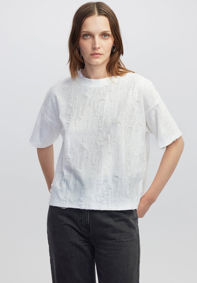 Witte kortearmige shirt met een gestructureerd oppervlak en subtiele parelaccenten. Comfortabele pasvorm, ronde halslijn en een zoom die aan de achterkant iets langer is.