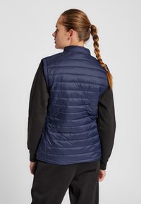 Mörkblå vadderad väst med horisontell sömnad, hög krage och ärmlös design. Modellen har en svart långärmad tröja under.