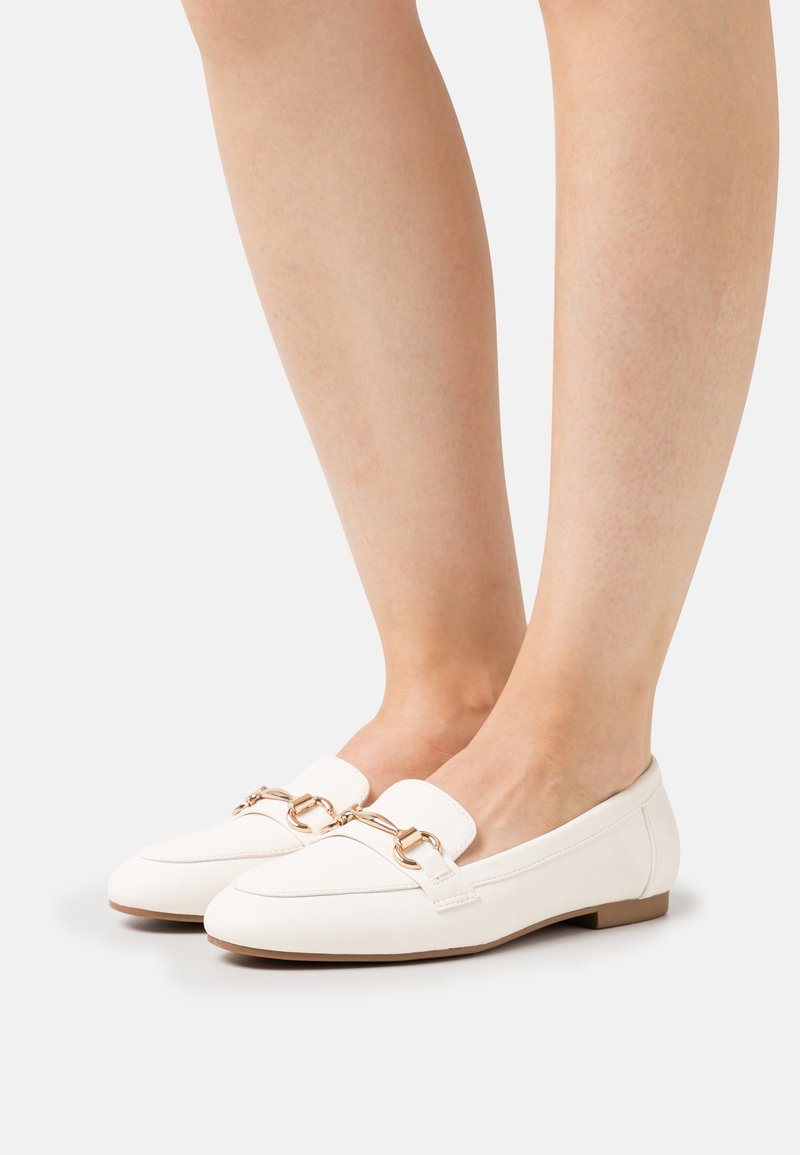 Des pieds chaussés de mocassins en cuir blanc avec un détail de mors doré et des semelles brunes sur un fond clair.
