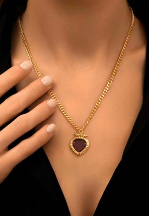 Collier chaîne en or avec un pendentif en forme de cœur en pierre précieuse violet foncé porté autour du cou d’une personne, main touchant délicatement la chaîne.
