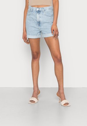 Femme portant un short en jean bleu clair retroussé et des sandales beiges à talons hauts avec des brides tressées, debout devant un fond uni.